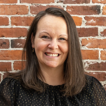 Smiling woman with shoulder-length brown hair against a brick wall.