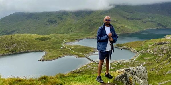 Yaya standing on top of a mountain with a lake in the background. He's holding his cane in his hands