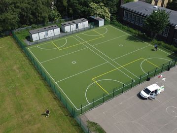 A fenced sports court with markings, surrounded by greenery and adjacent to a parking lot.