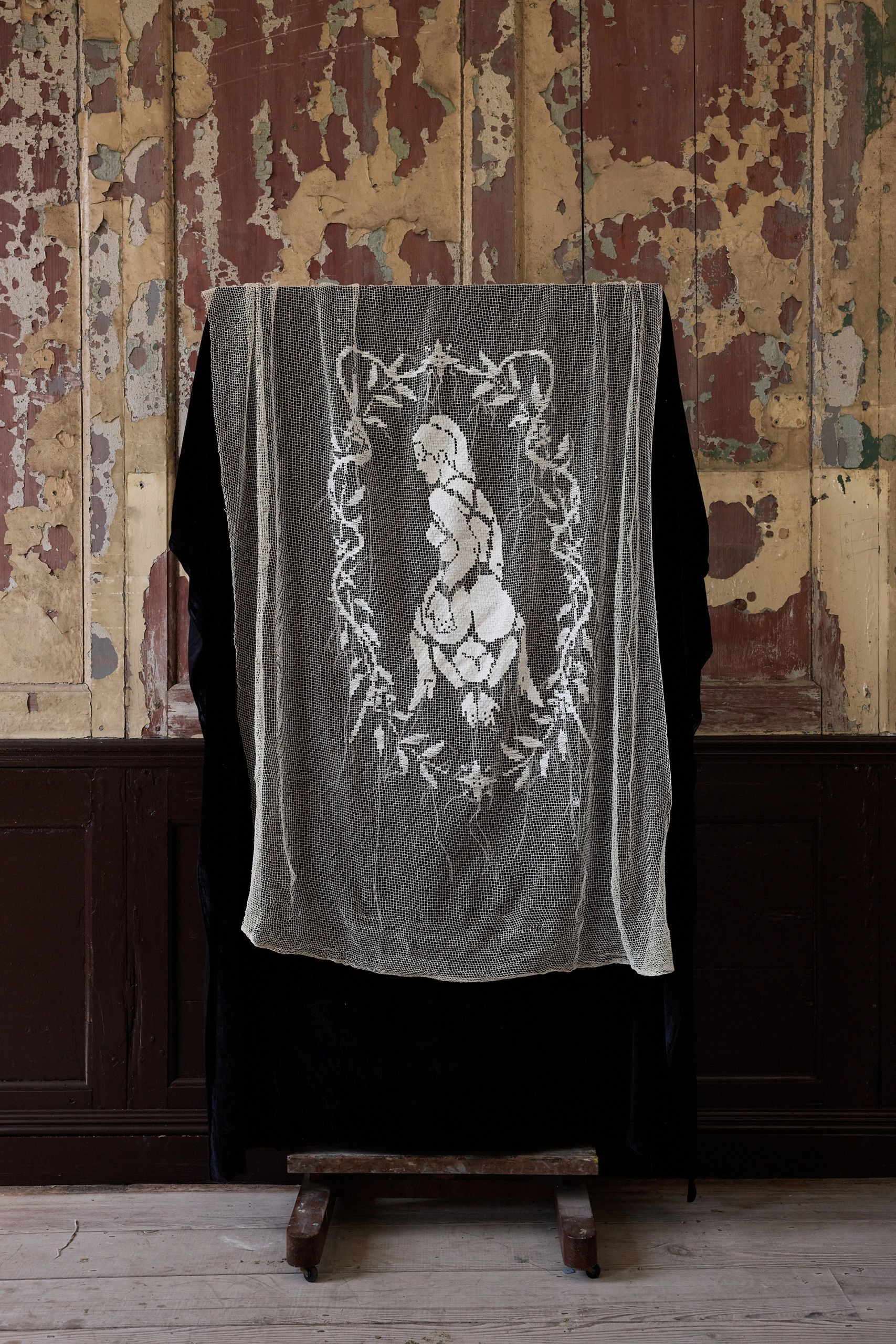 Lace curtain with a female figure design in front of a peeling wall.
