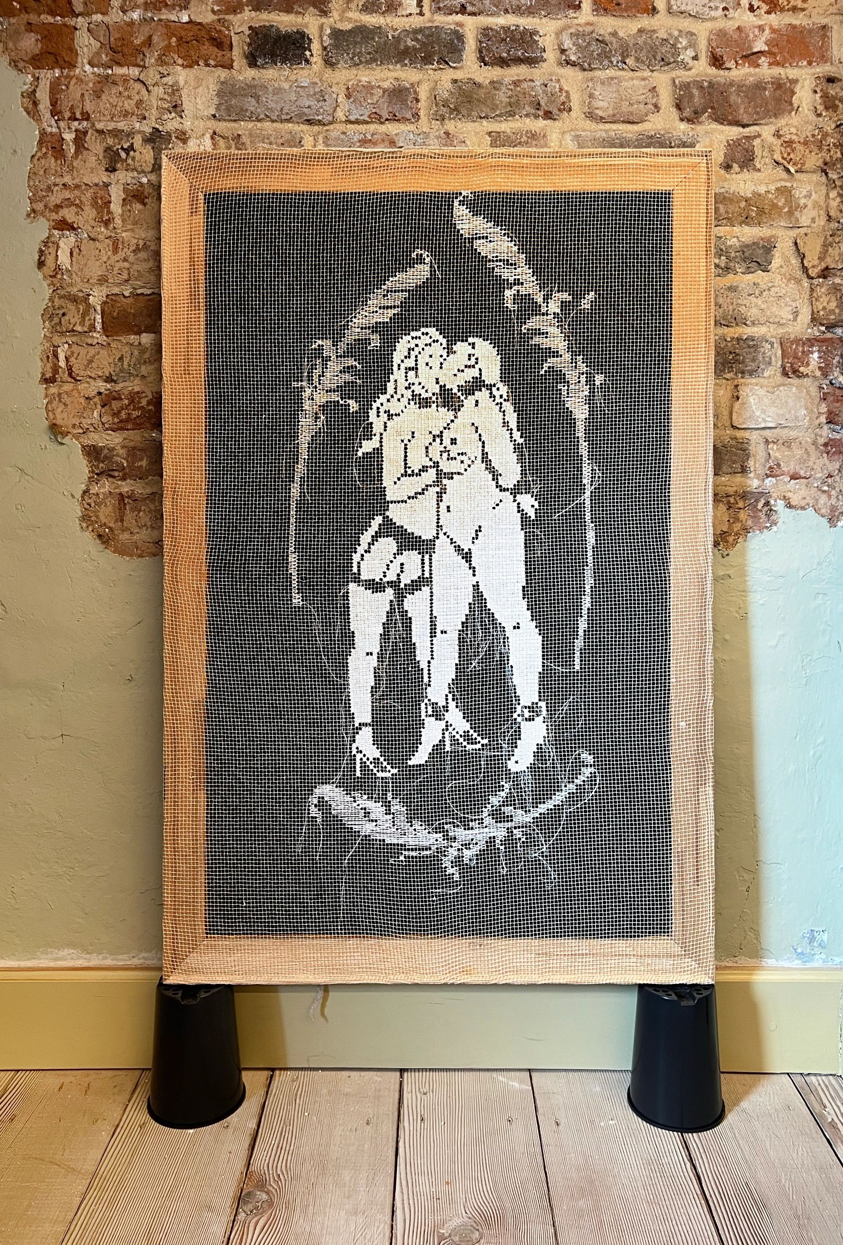 Cross-stitched artwork of two women kissing on a brick wall.