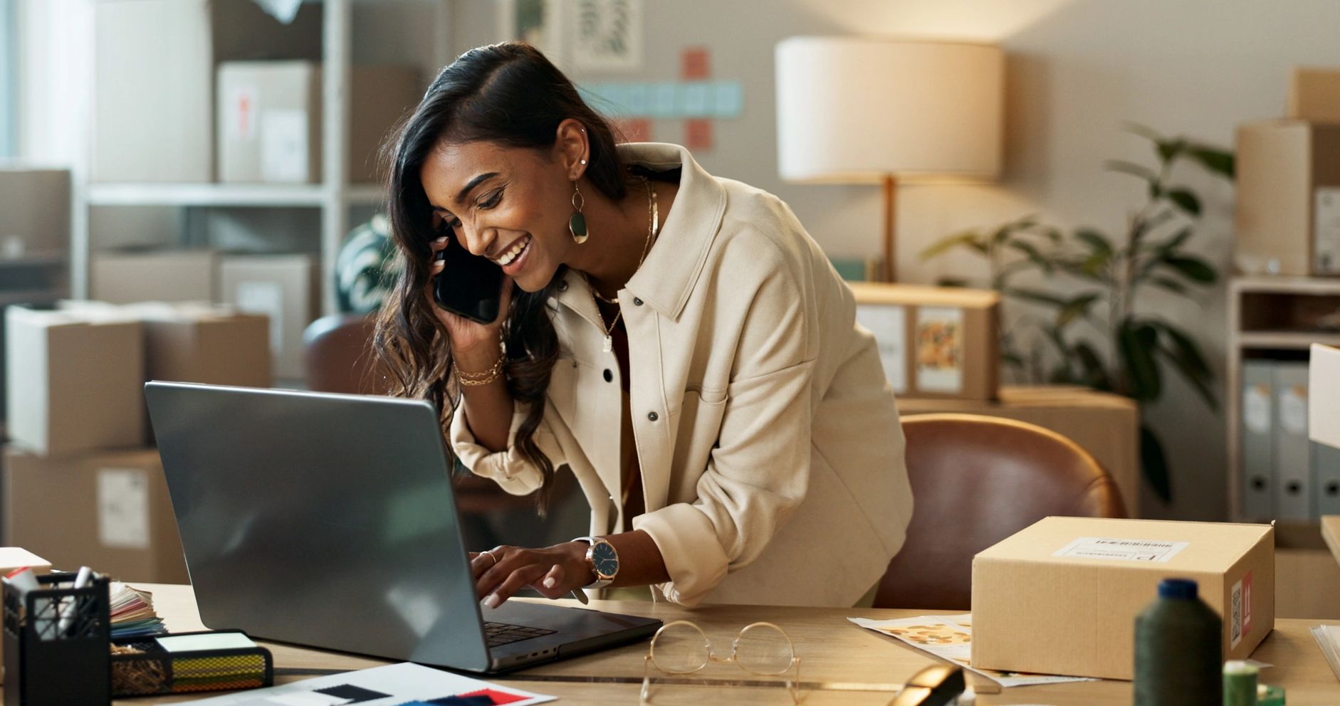 Smiling woman talks on phone while working on laptop in a cozy office.