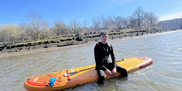 A young man in a wetsuit sitting on a paddleboard in calm water near a shore.