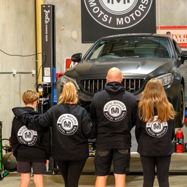 Family wearing matching Motsi Motors hoodies in a garage with a car.