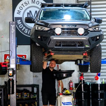 Mechanic working under lifted Isuzu truck in a garage.