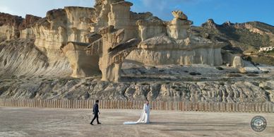 A couple in wedding attire walking near unique rock formations under a clear blue sky.