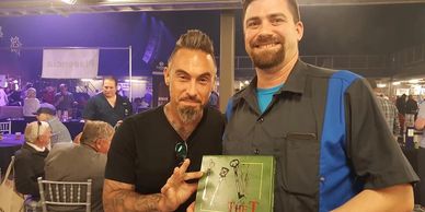 Two men pose with a signed album at an event with seated attendees in the background.