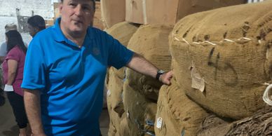 Man in blue shirt with cigar next to stacked burlap sacks in warehouse.