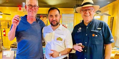 Three men smiling and holding cigars in a cozy indoor setting.
