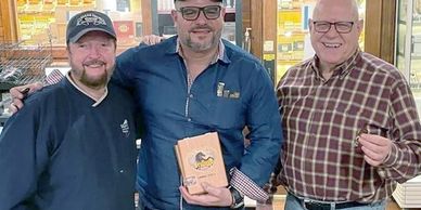 Three men smiling, one holding a wooden box, standing indoors.