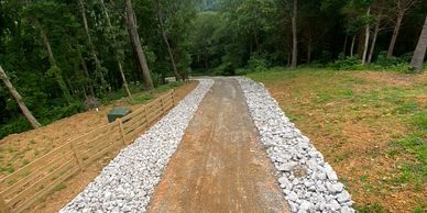A gravel pathway bordered by white rocks and a wooden fence through a wooded area.