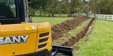 Yellow excavator digging a long trench in grassy field near white fence.