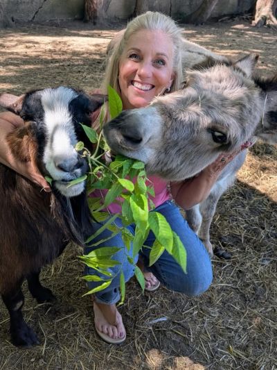 Woman smiling with a goat and donkey eating leaves.