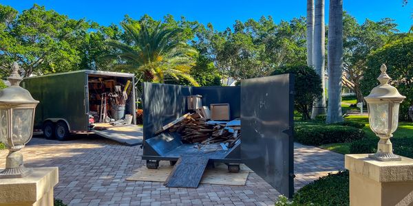 Open dumpster and trailer with debris in a sunny driveway.