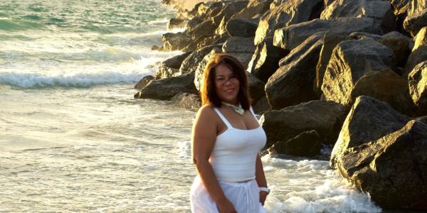 Woman in white dress standing by rocky shore at sunset.