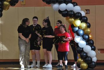 A group of cheerleaders and a woman standing under a balloon arch indoors.
