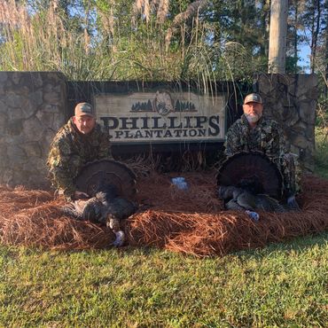 Two hunters in camouflage holding turkeys in front of Phillips Plantation sign.