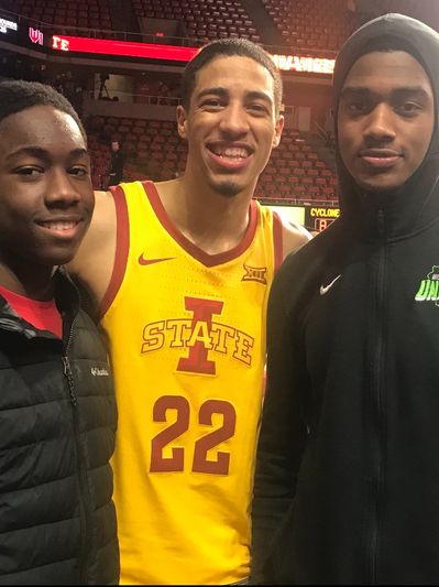Three young men posing in a sports arena, one in a yellow basketball jersey.
