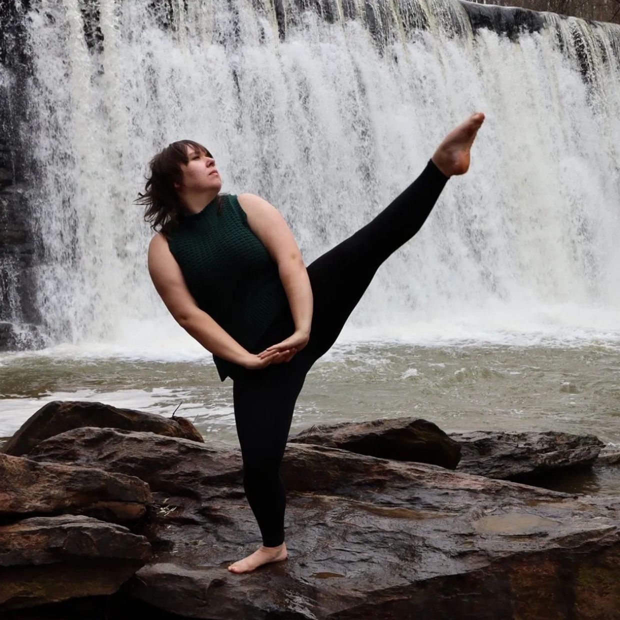Cecilia stands in front of a waterfall on one leg with her other leg extended in the air