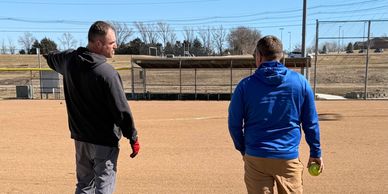 Two men stand on a baseball field, one pointing while the other holds a softball.