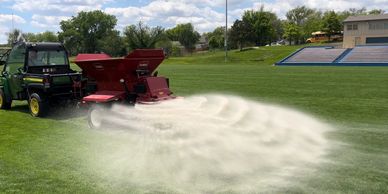 A vehicle spreads white powder on a green sports field under a blue sky.