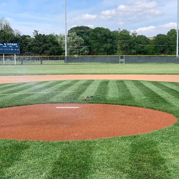 Baseball field with a sprinkler on the pitcher's mound and scoreboard in the background.