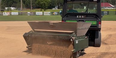 A vehicle spreads sand on a baseball field with a Turfco spreader.