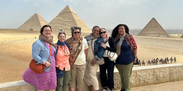 Group of six friends posing with the pyramids of Giza in the background under a cloudy sky.