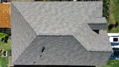 Aerial view of a gray shingled house roof with a chimney and surrounding greenery.
