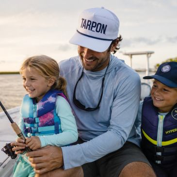 A man helps two kids fish on a boat, all smiling and wearing life jackets.
