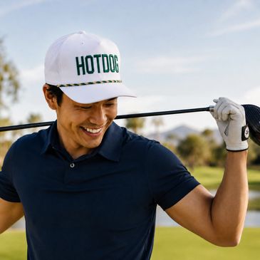 Smiling man in a white 'HOTDOG' cap holding a golf club on a sunny golf course.