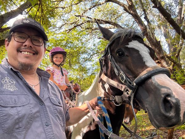 Man and child smiling with a horse in a sunny wooded area.