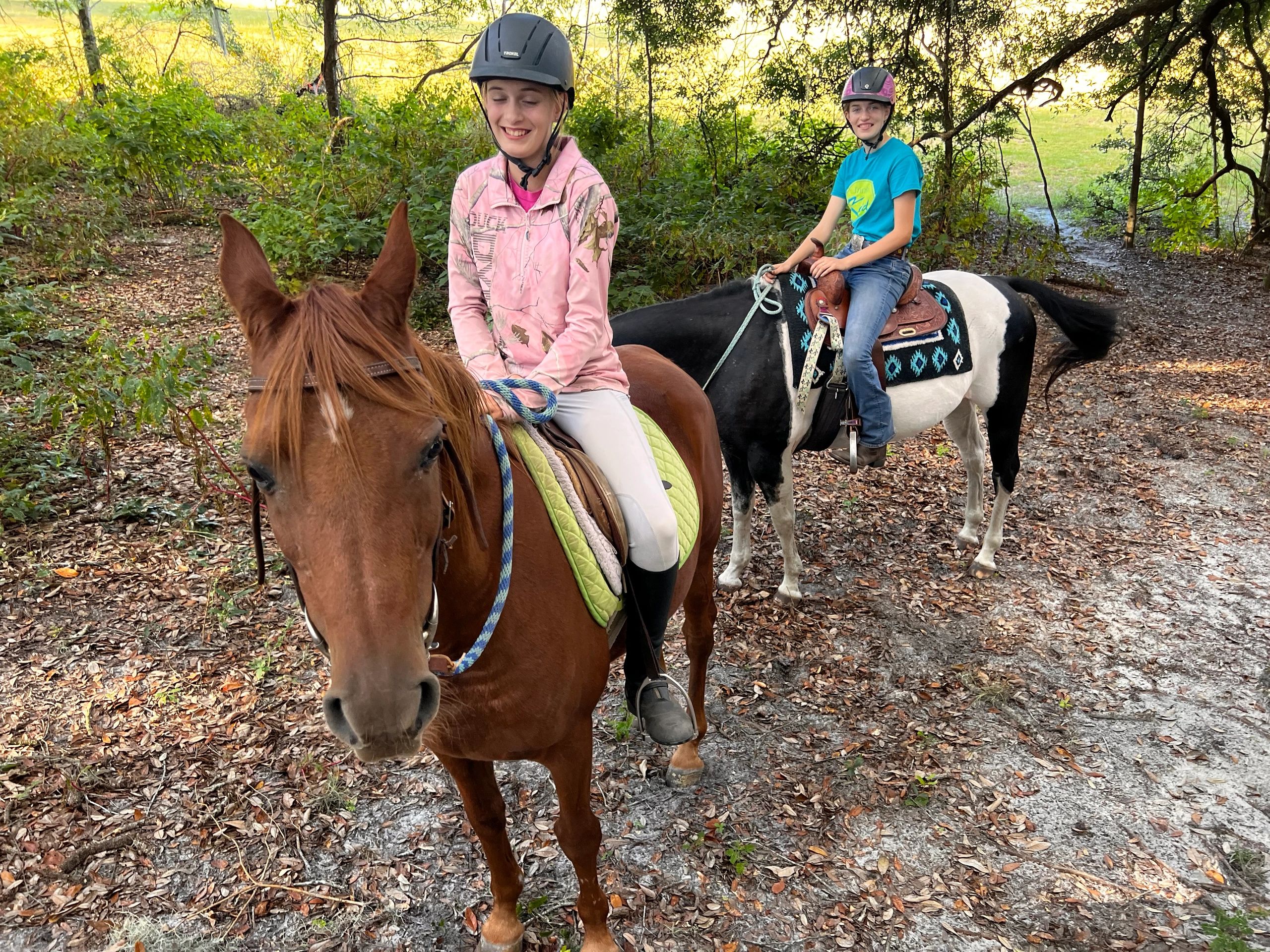 Two people riding horses on a leafy trail wearing helmets.