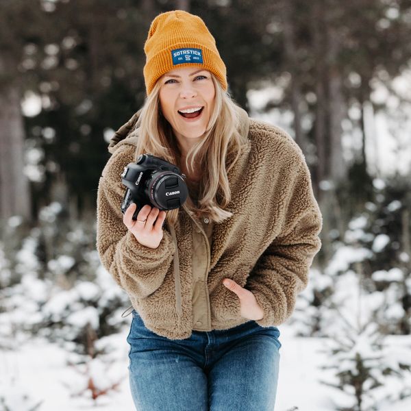 Woman in winter clothes holding a camera and smiling outdoors in the snow.