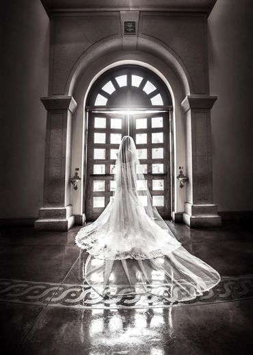Bride in a flowing gown stands before an ornate arched doorway with sunlight streaming in.