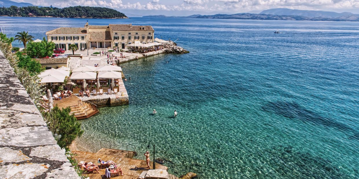 Sunbathers relax by a historic waterfront with clear turquoise waters and distant islands.