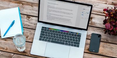Laptop with document on screen, notebook, pen, glass of water, and phone on wooden table.