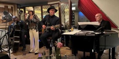 A lively band performs with guitar, piano, and vocals in a cozy indoor setting.