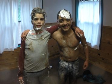 Two young men covered in foam and mud, smiling indoors.