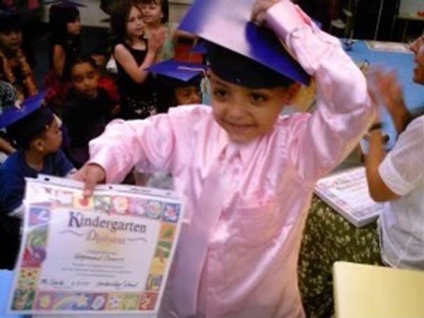 A young boy proudly holds his kindergarten diploma wearing a graduation cap.