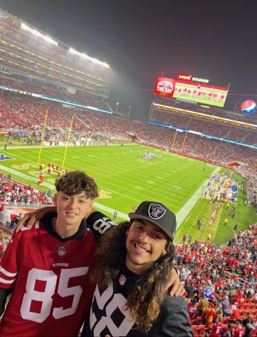 Two friends smiling at a football stadium during a night game.