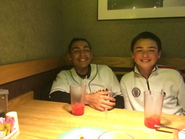 Two boys smiling at a restaurant table with drinks in front of them.