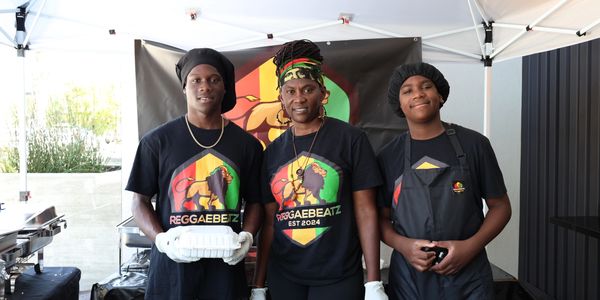 Three people wearing Reggaebeatz shirts stand under a tent, smiling.