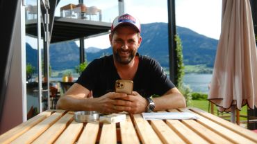 Mann mit Baseballkappe sitzt draußen und lächelt mit Handy in der Hand.