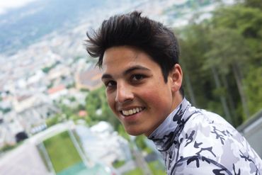 Junger Mann mit schwarzem Haar lächelt in Kamera vor Berglandschaft.