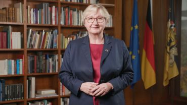 Frau steht vor Bücherregal und Flaggen.