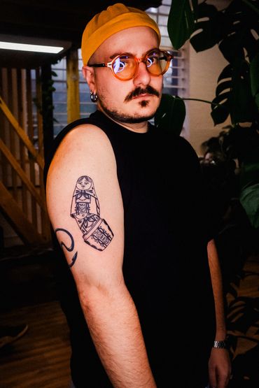 Man with a yellow beanie and orange glasses showing a detailed tattoo on his arm.