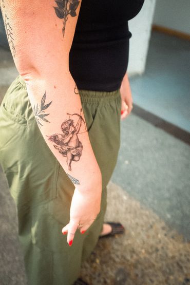 Tattoo of a cherub with a bow and arrow on a forearm with other tattoos.