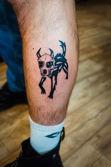 Black ink tattoo of a creature with a skull-like face on a person's leg.