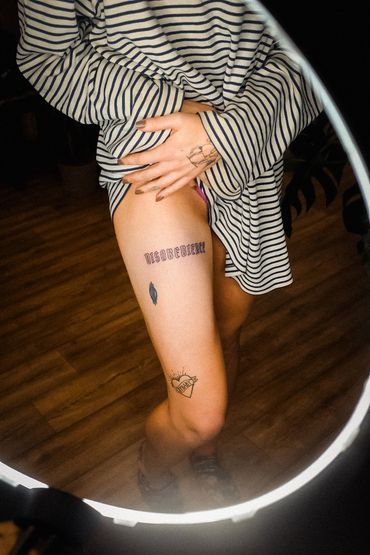 Person showing thigh tattoos through a striped shirt, standing on wooden floor.